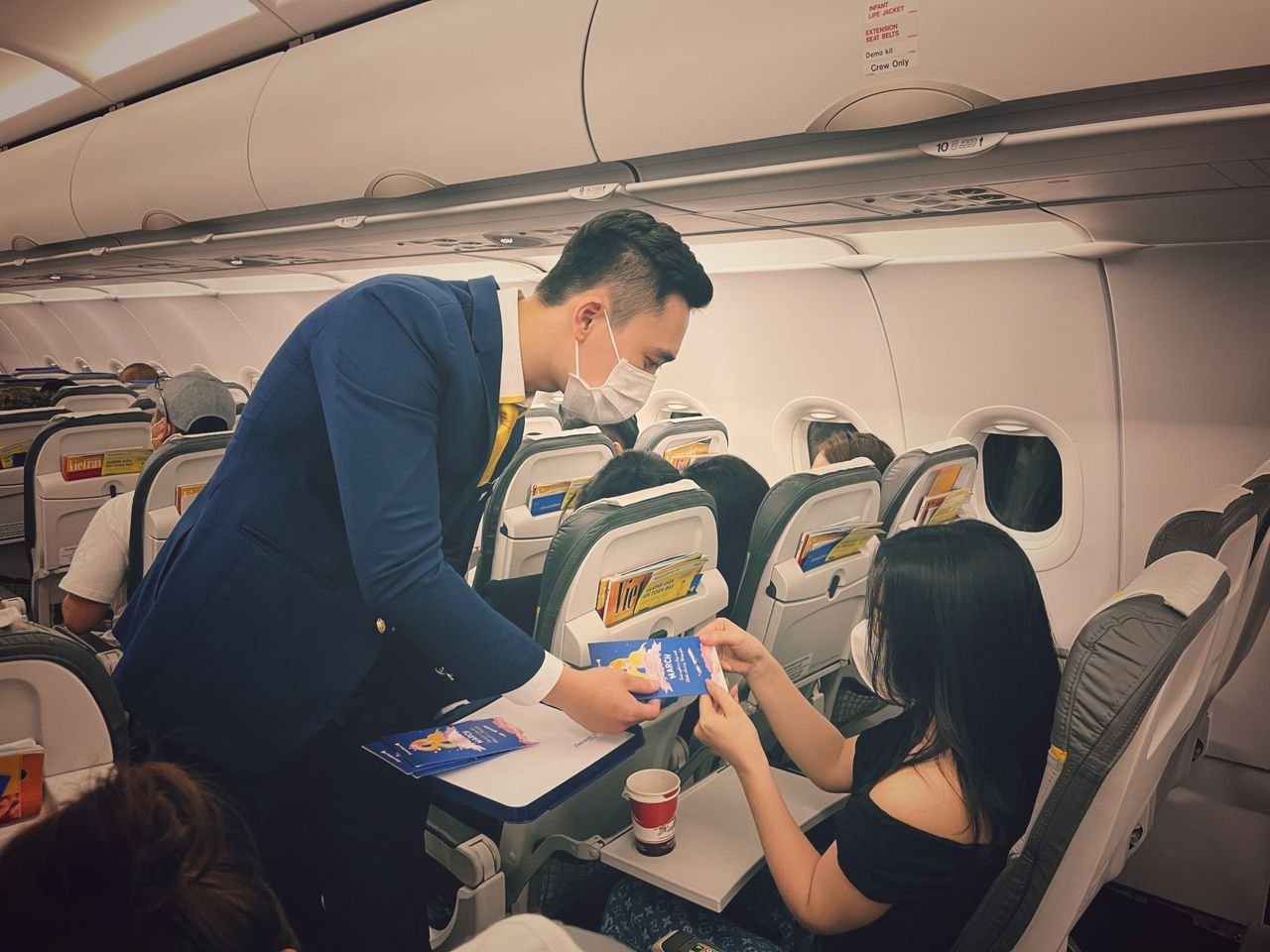 Flight attendant in mask serving passengers on an airplane, ensuring safety and comfort.